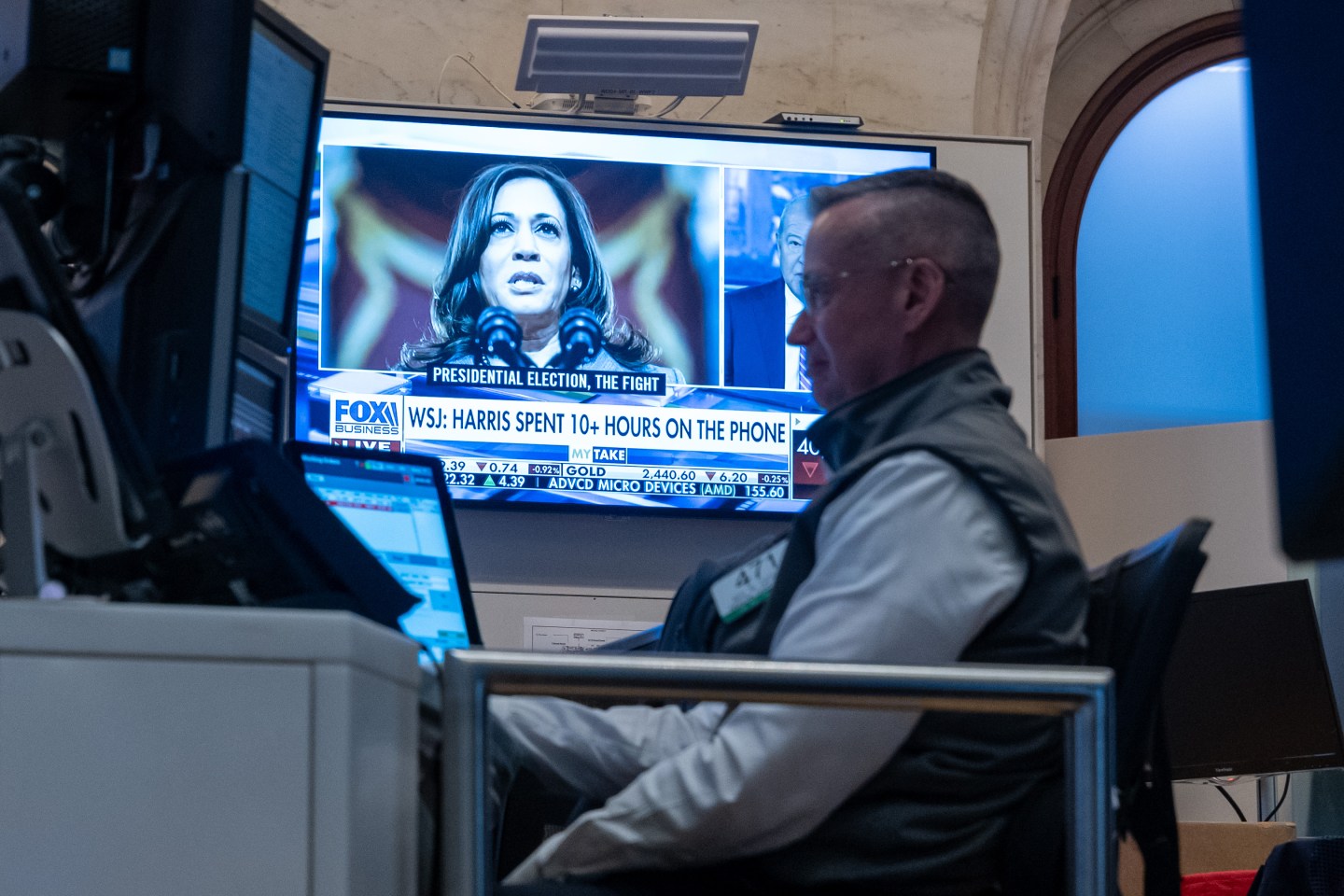 A trader on the floor of the New York Stock Exchange works while a TV shows footage of Kamala Harris in the background.