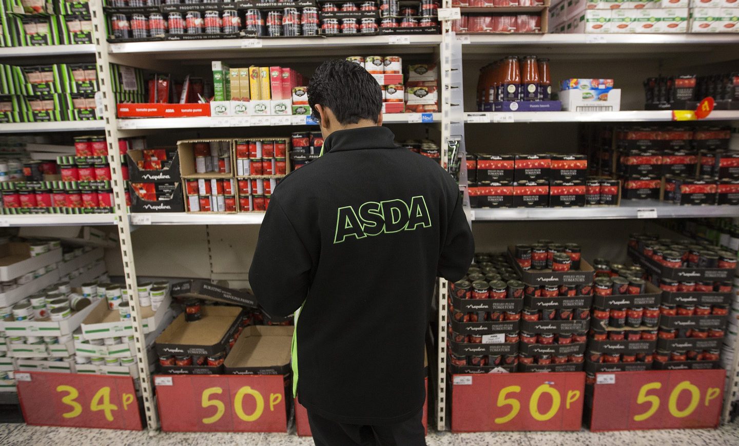 FILE: An Asda logo sits on an employee's jacket as he works near a display of tinned food inside an Asda supermarket, the U.K. retail arm of Wal-Mart Stores Inc., in Watford, U.K., on Thursday, Oct. 17, 2013. U.K. grocer J Sainsbury Plc plans to buy Walmart Inc.s Asda in a 7.3 billion-pound ($10 billion) deal that would transform the countrys supermarket industry and leave the U.S. retailer as the combined companys biggest shareholder. Our editors select the best archive images of the two supermarket chains.