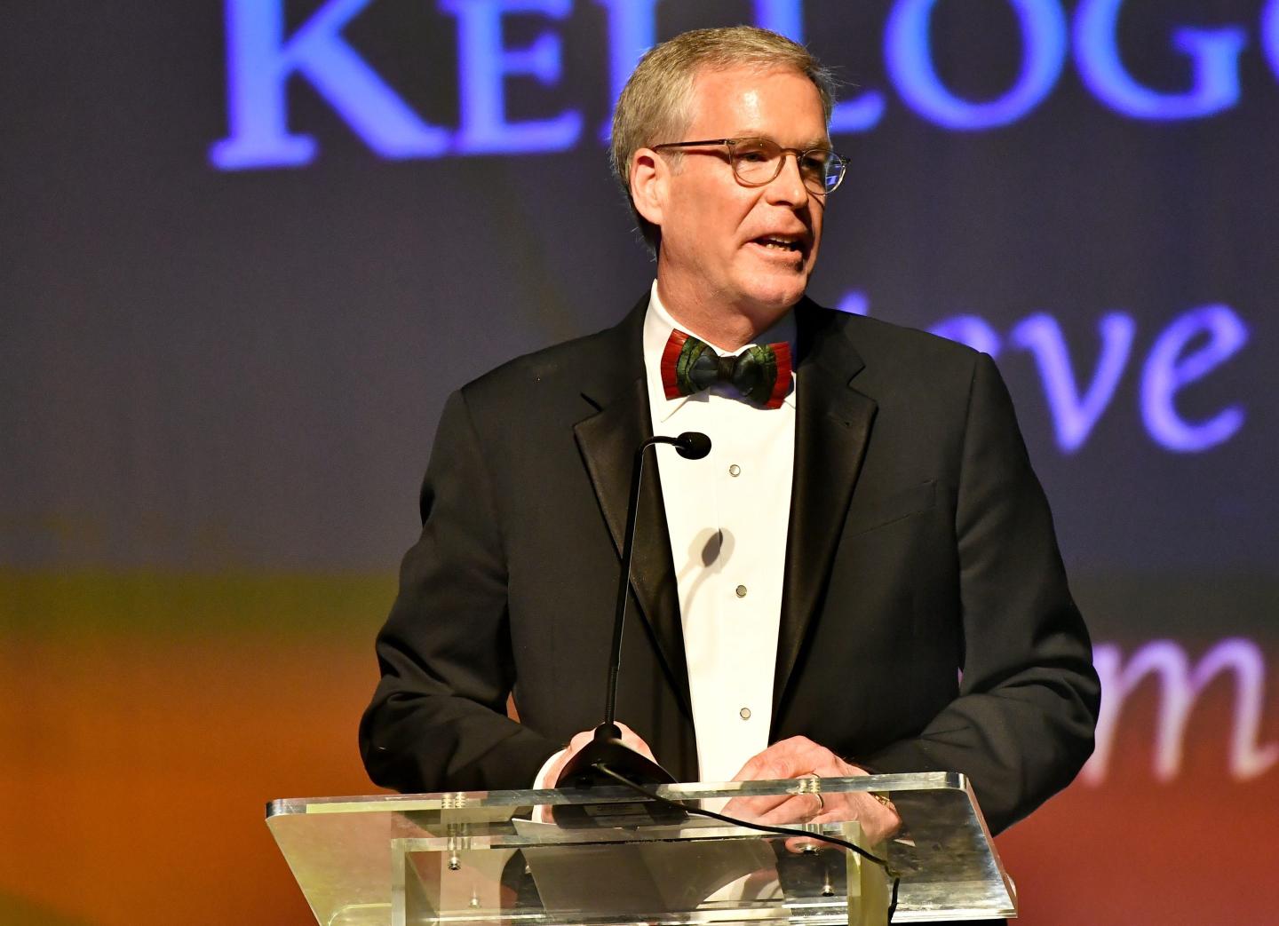 Steve Cahillane wearing a suit and bowtie speaks behind a glass podium.