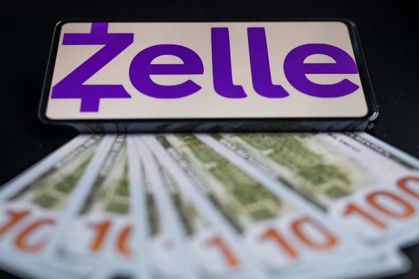 A phone displaying the Zelle logo at the top of an image with one hundred dollar bills splayed out in front of it.