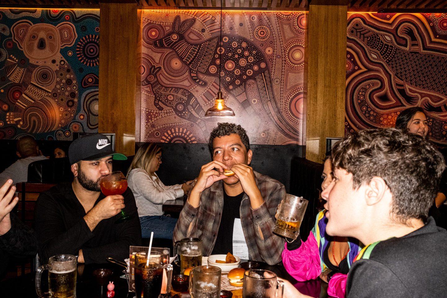 A man in center of the photo puts a slider in his mouth as people at his table sip drinks and talk during dinner. In the background, there are mosaics of Australian animals like kangaroos and koalas.