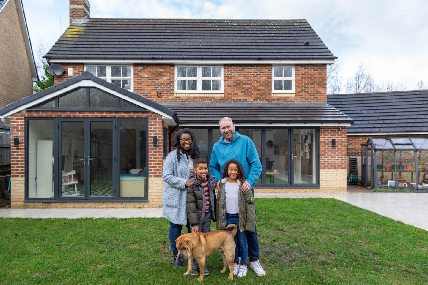 A family standing proudly in front of the home in the garden. They are all looking at the camera with positive emotion. The dog is walking around their legs.