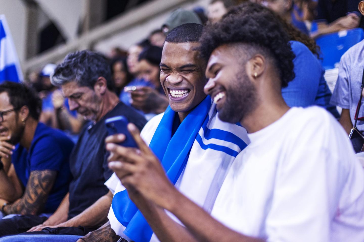 Two young men sit in the stands at a sports event and smile while looking at a phone