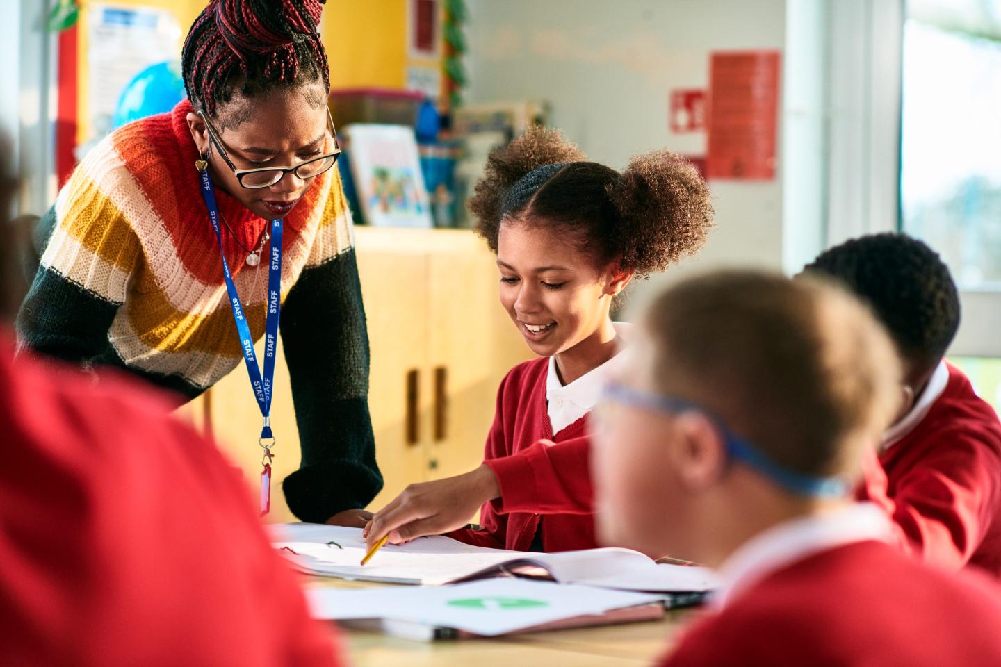 Cheerful elementary school girl showing her book to her teacher who is leaning over the desk and concentrating