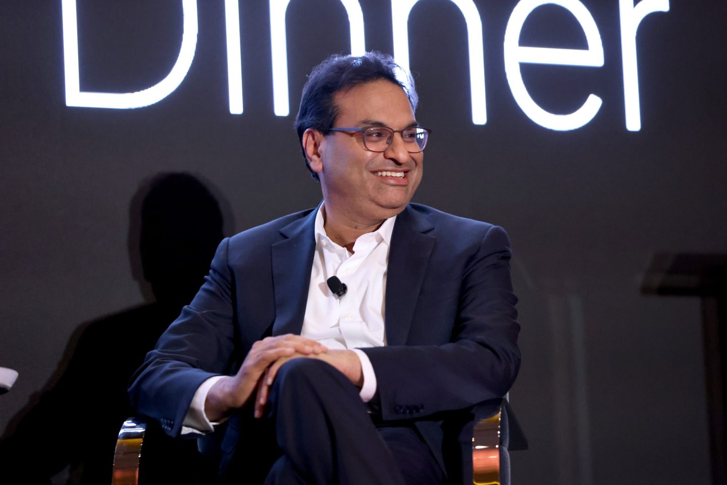 Laxman Narasimhan, dressed in a light shirt and suit, sits with his hands on his knee and looks out toward the crowd, smiling, at a conference.