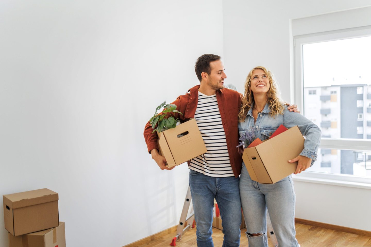 A man and a woman holding moving boxes stand in an empty room
