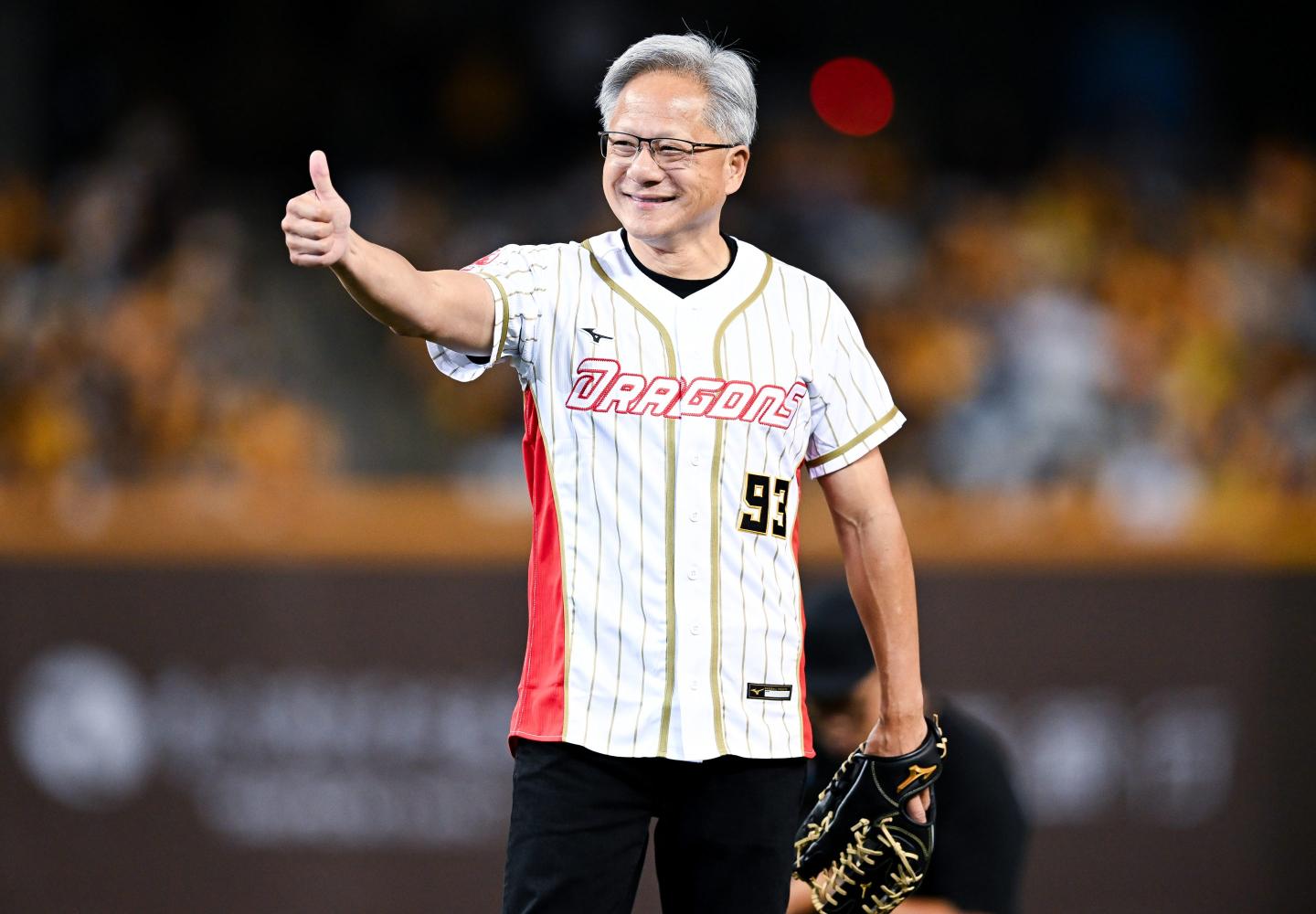 Jensen Huang, baseball glove in hand and wearing a baseball jersey, gives a thumbs up.