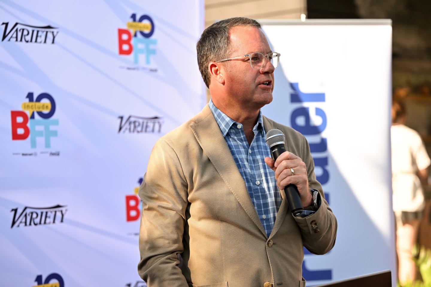 Doug McMillion wears and tan suit and speaks into a microphone.