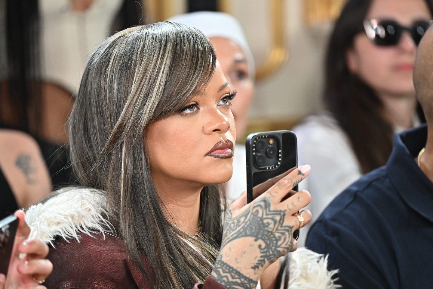 Pop star Rihanna, dressed in a red jacket, keeps a straight face as she holds her phone in the air with her right hand, presumably recording a performance.