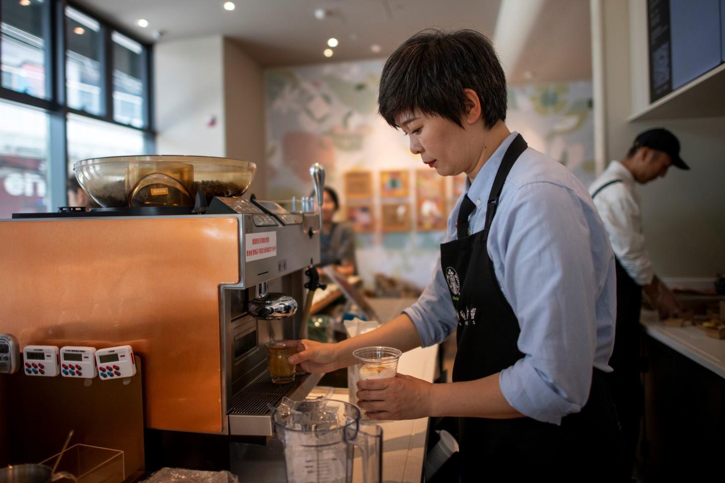 A Starbucks barista in China stands if front of a large coffee machine, with a drink in her left hand and another class under the machine.