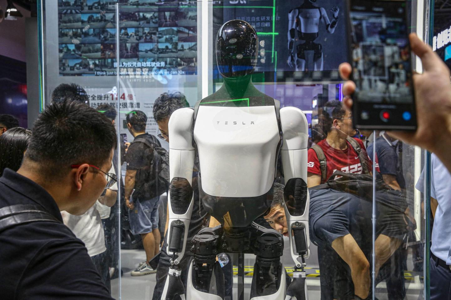 People look at an Optimus bot, which is standing in a clear glass box.