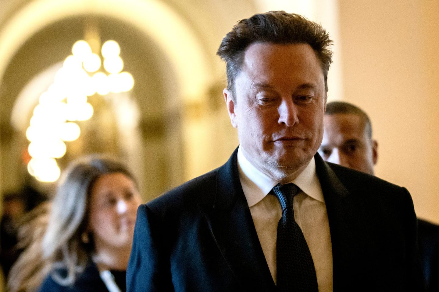 Elon Musk wears a suit and walks down a hallway.