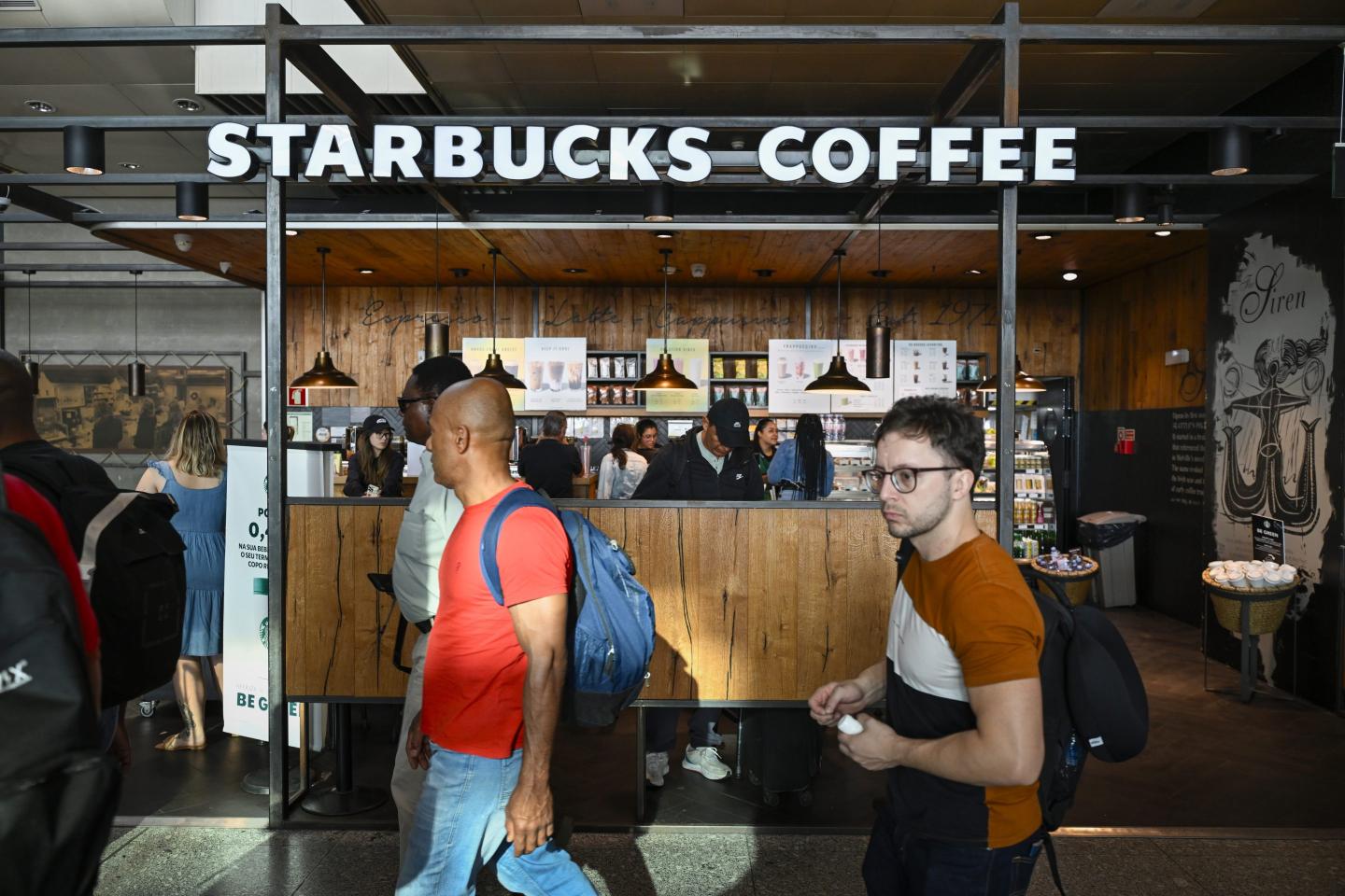 Tourists, including three men in the center of the image, walk past a small Starbucks cafe in a Lisbon airport.