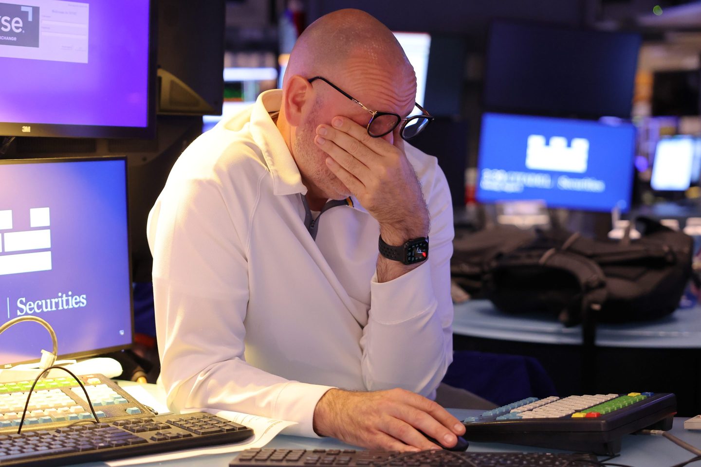 A man in a white sweater on the floor of the New York Stock Exchange puts his left hand over his face, pushing his glasses above his eyes. His right hand remains on his mouse amidst a background of many monitors and keyboards.