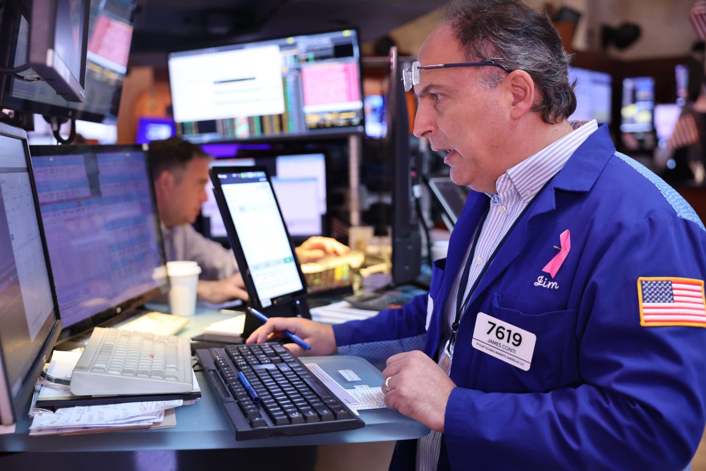 A man on the floor of the New York Stock Exchange looks intently at his screen with his mouth open and glasses pushed onto his forehead.
