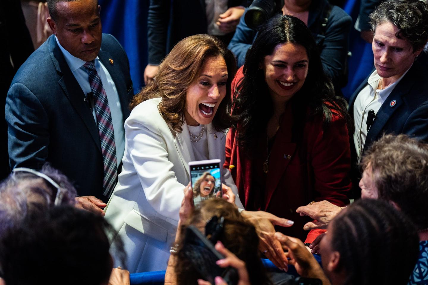 Kamala Harris wearing a white suit, is enthusiastically greeting and reaching out to a crowd of supporters while smiling widely. She is surrounded by people, including a security detail and another woman in a red jacket. Many in the crowd are capturing the moment on their phones. The atmosphere is energetic and joyful.