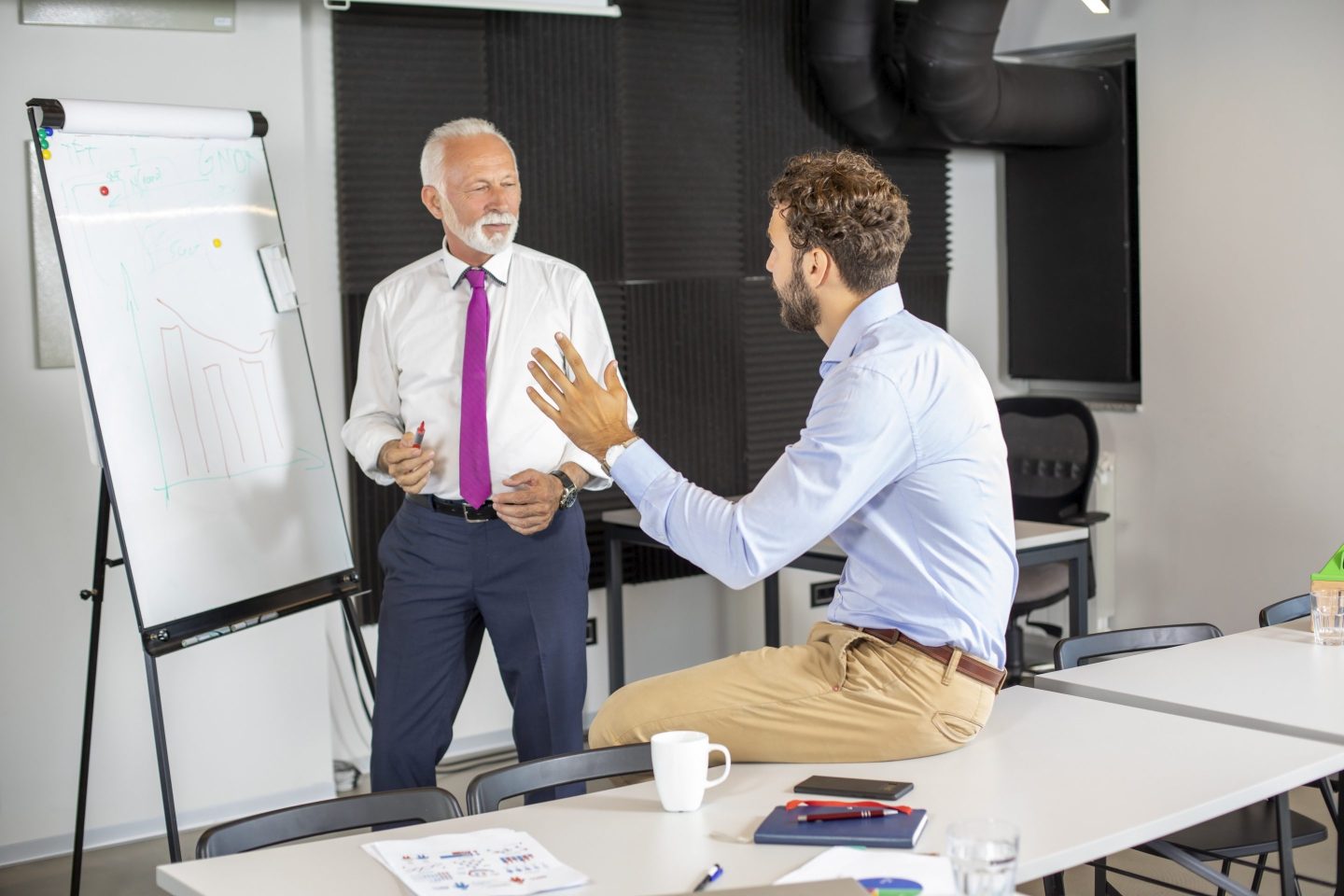 Business person next to a presentation board explains a concept to a younger business person.