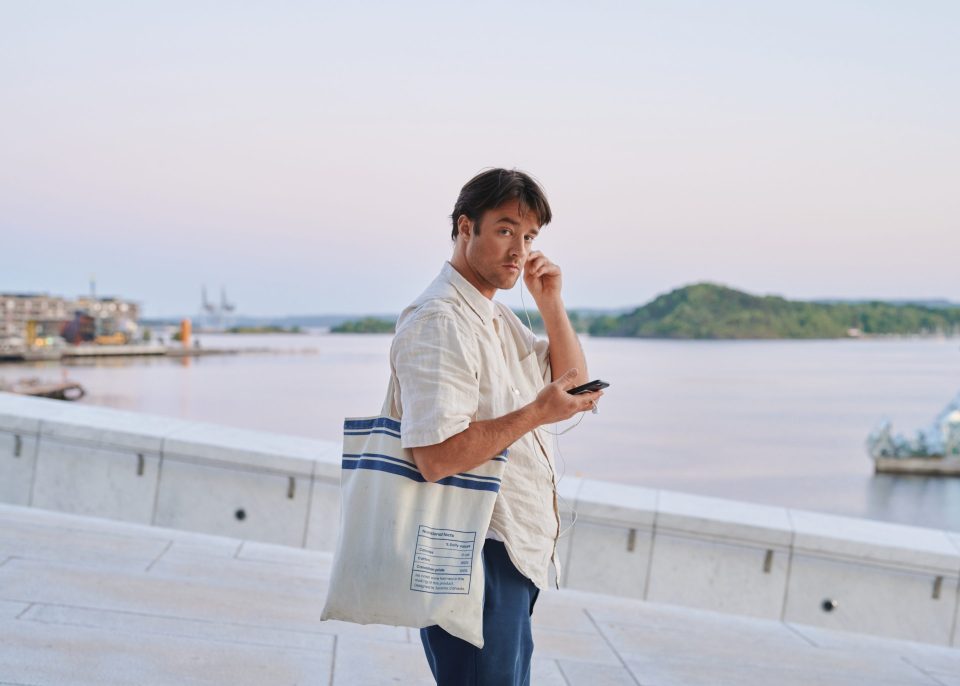 A man with a tote bag puts an earphone in his ear and looks at the camera.