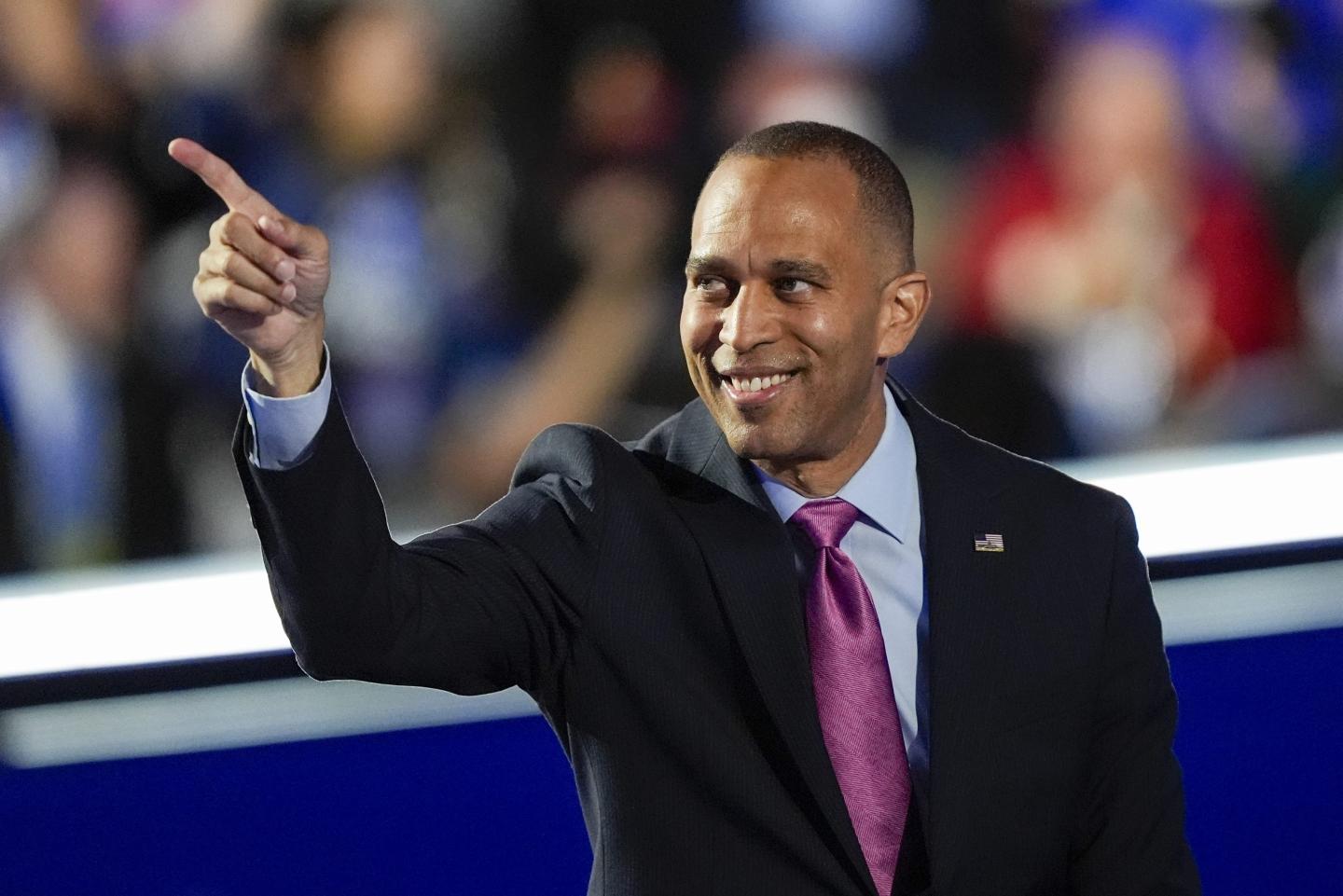 Rep. Hakeem Jeffries stands in front of a blurred crowed, pointing into the audience and smiling. He wears a black suit with a light blue shirt and a magenta tie.