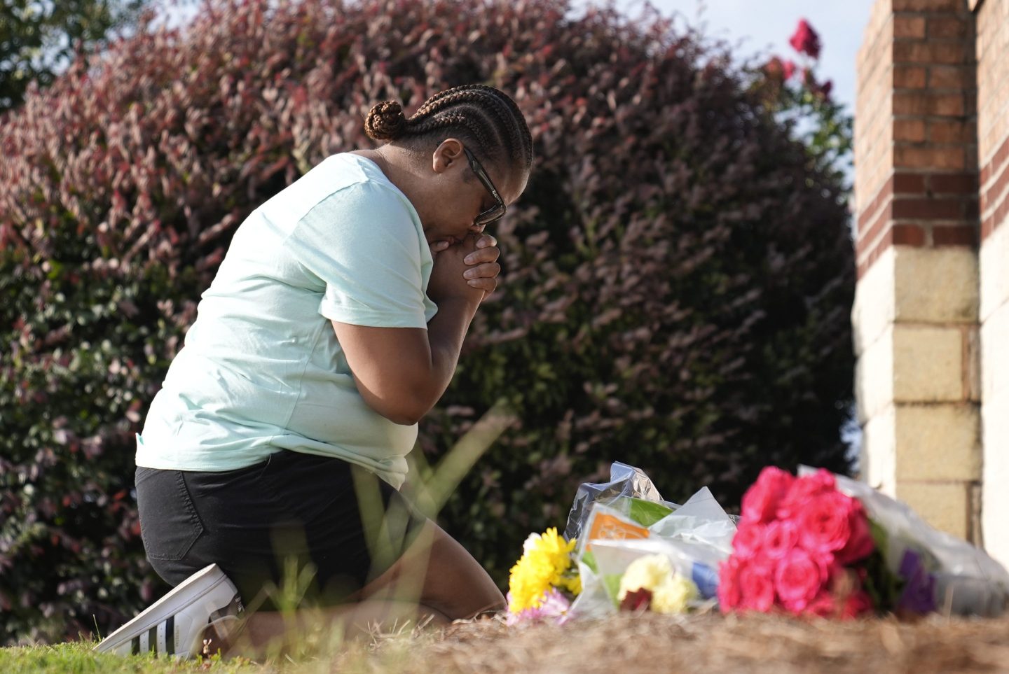 A woman kneels on the ground in reflection in front of a small memorial of flowers. She appears to be mourning, with her hands clasped and head bowed.