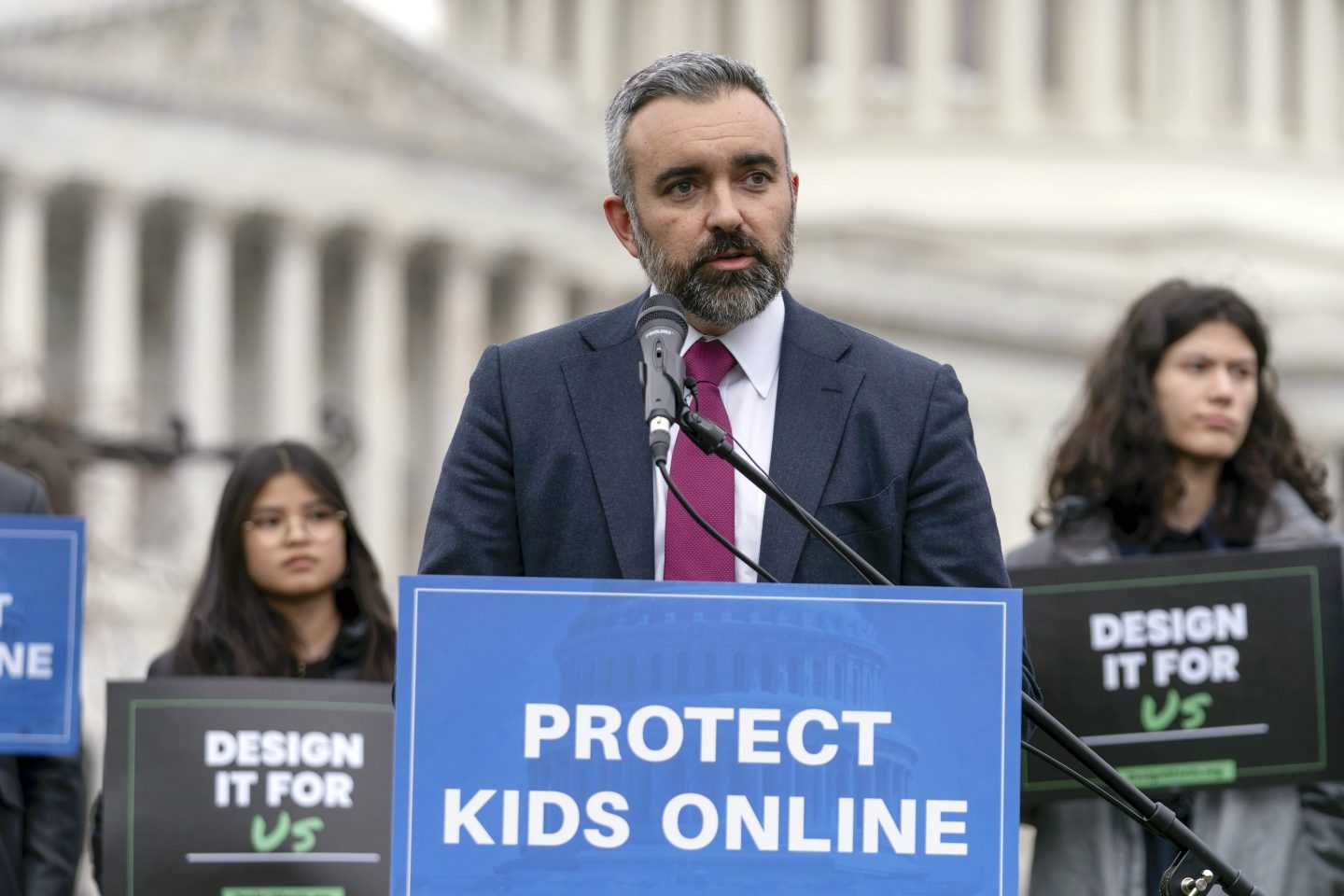 New Mexico Attorney General Raúl Torrez speaks in front of a podium with a blue sign reading "Protect Kids Online." In the background is the blurred capitol building.