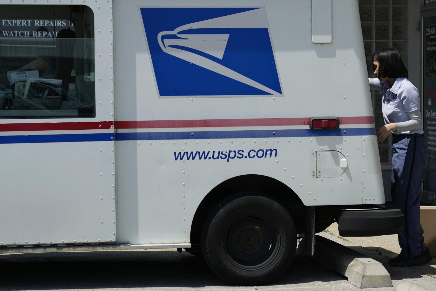 A USPS truck is parked on the sidewalk on a sunny day. The USPS logo is clearly visible and a mail carrier is seen at the back of the truck unloading parcels.