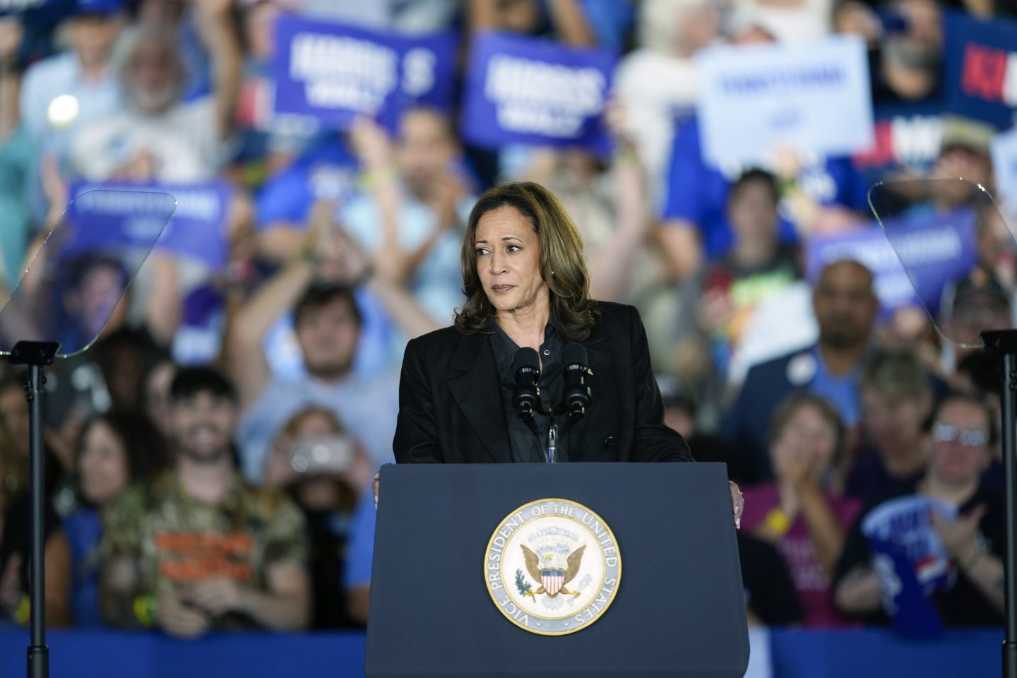 Kamala Harris is seen behind a Vice President podium as a blurred crowd of supporters in the background hold supportive signs.