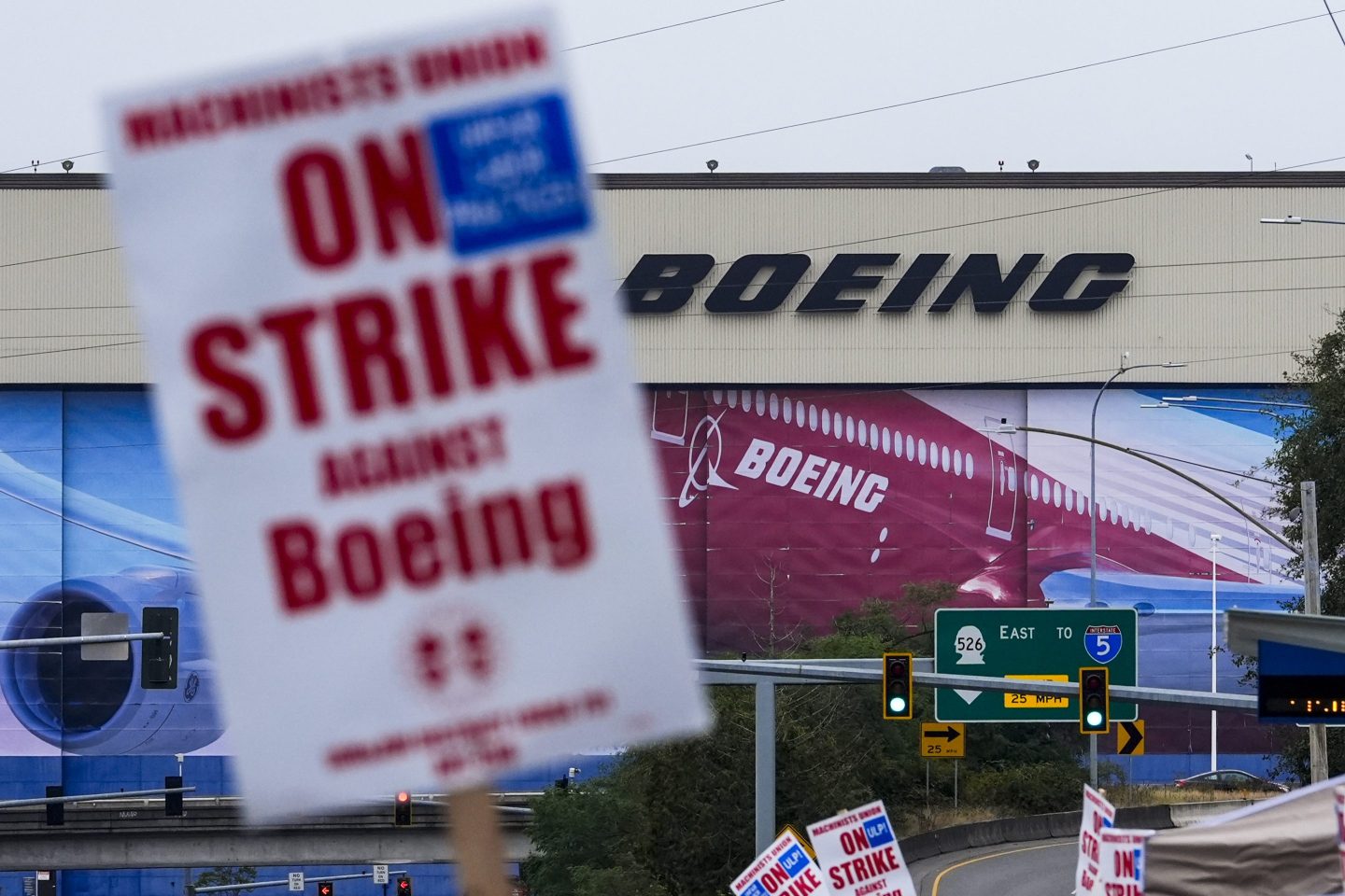 The image shows a protest scene in front of a Boeing facility, with large Boeing logos visible on the building. In the foreground, multiple signs held by union members are visible, with one prominently displaying “On Strike Against Boeing” in bold red letters. The strike appears to be organized by the Machinists Union, with additional signs in the background. The industrial setting and signage emphasize the labor protest against the aerospace company.