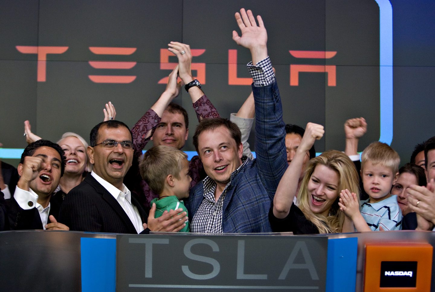 Elon Musk, surrounded by a crowd of people on the stage for the Nasdaq opening bell ceremony, holds his son and waves his other hand high in the air.
