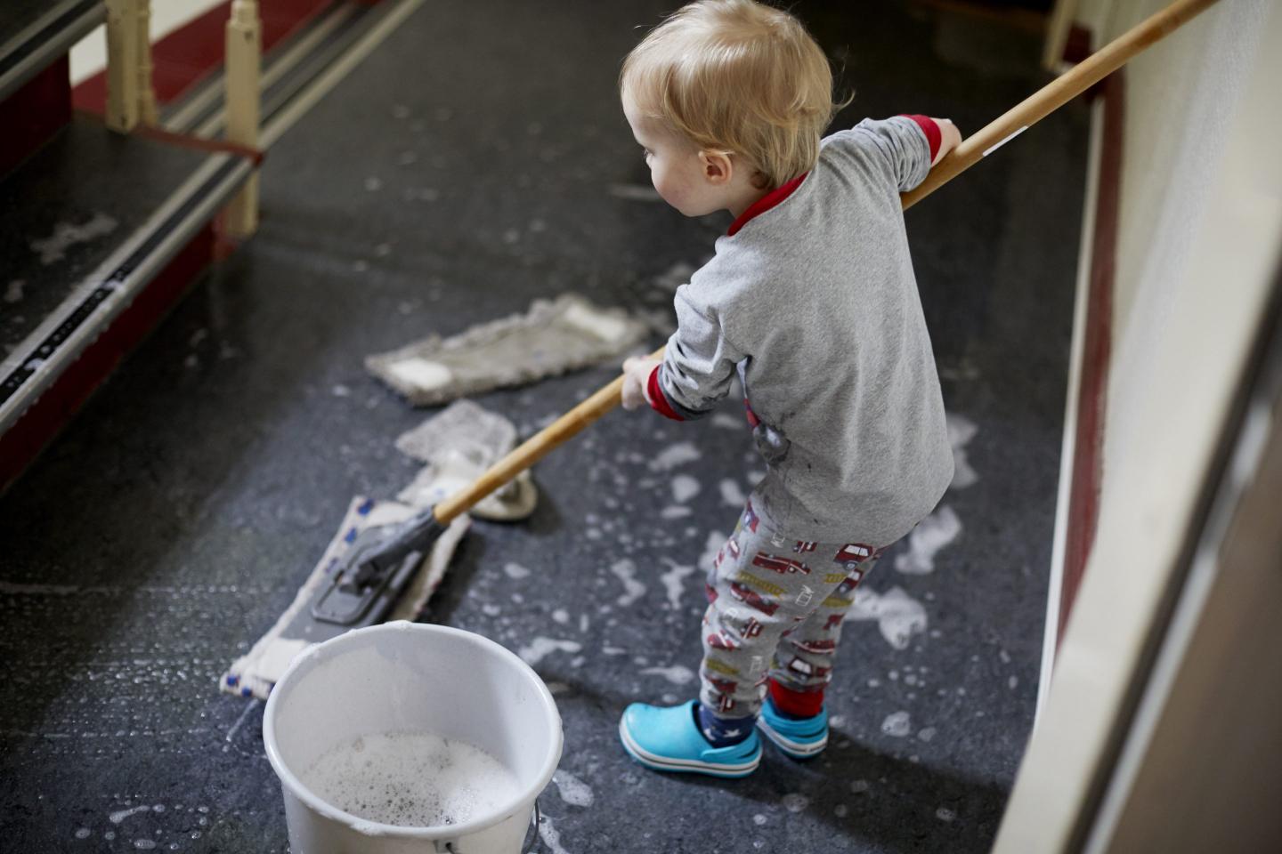 A toddler in pajamas mops the floor of a hallway. Soap is visible on the floor and the child looks away from the camera.