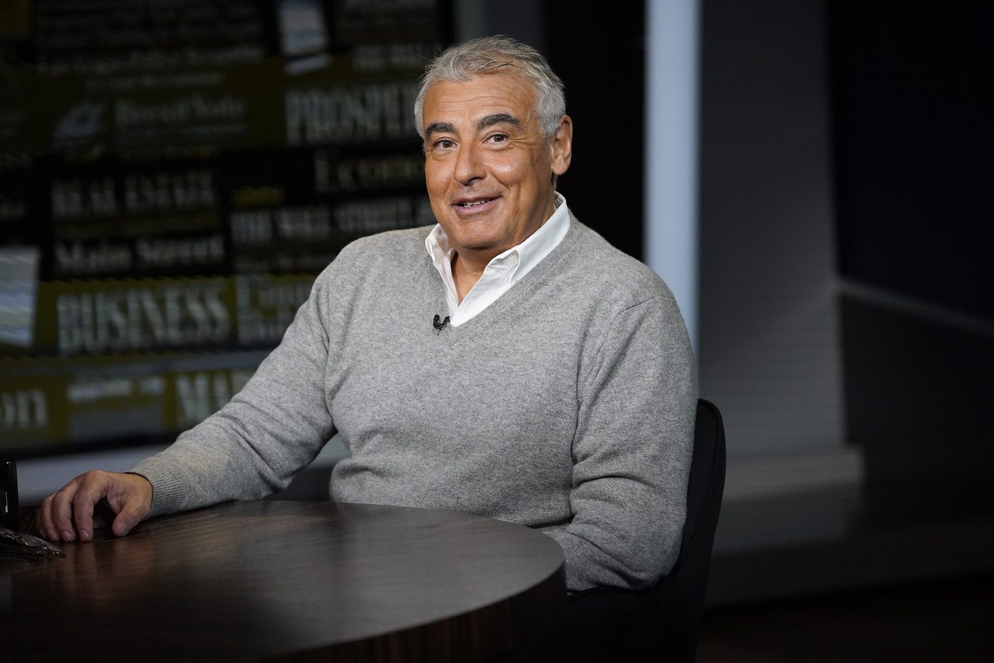 Marc Lasry sits at a desk in a gray sweater with a white suit collar layered underneath. He is smiling looking directly at the camera.