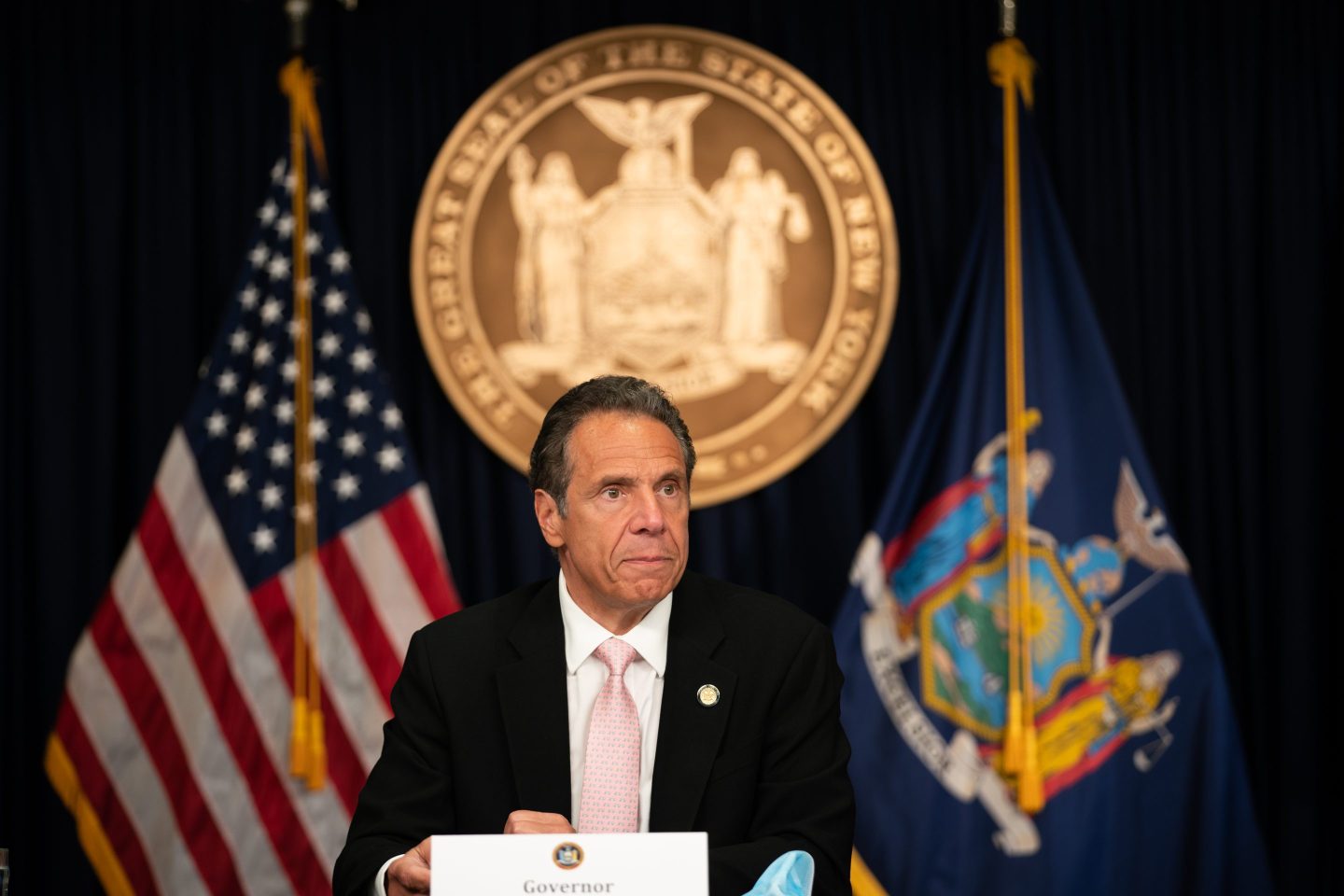 Former New York Governor Andrew Cuomo sits at a desk during a press conference, with the Great Seal of the State of New York and American flags displayed behind him. Cuomo, wearing a black suit, white shirt, and pink tie, has a serious expression on his face as he looks slightly to the side. The setting is official, reflecting his role as governor at the time.