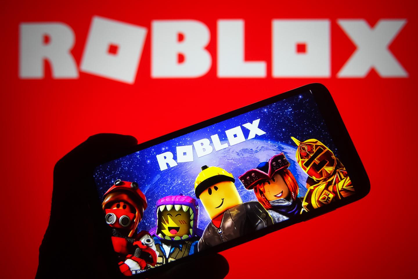 A mobile phone is running the Roblox game app, showing five avatars beneath the Roblox logo.