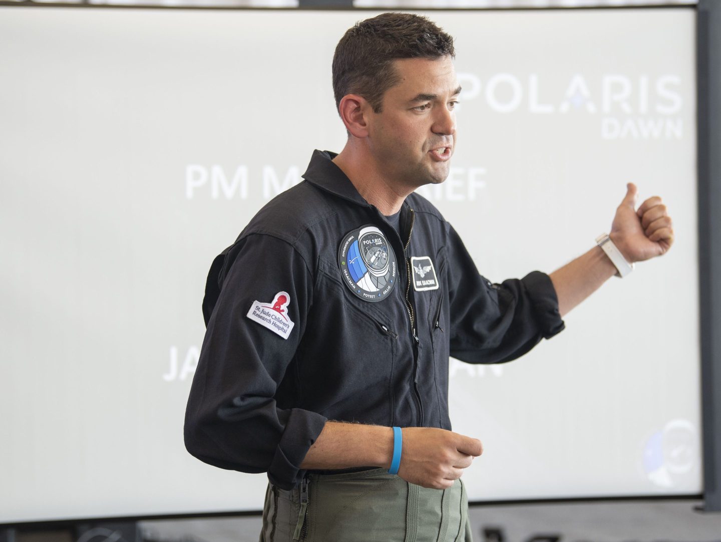 Billionaire Jared Isaacman, dressed in a black flight suit adorned with mission patches, stands in front of a projection screen giving a presentation. He gestures with his right hand, appearing mid-speech, preparing for space travel as part of the Polaris Dawn mission. His flight suit features patches, including one for St. Jude Children's Research Hospital. The background shows the partially visible Polaris Dawn logo on the screen.