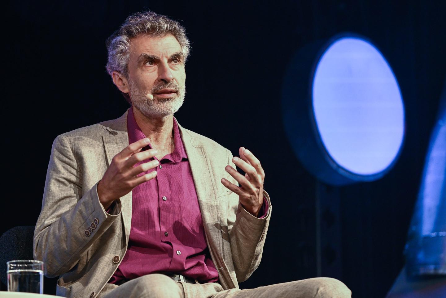 Yoshua Bengio, professor at the Montreal Institute for Learning Algorithms, is seen at a lecture event wearing a tan suit with a maroon dress shirt. His mouth is slightly open as he speaks and he is wearing a microphone. His hands are raised as he gestures while speaking on AI.