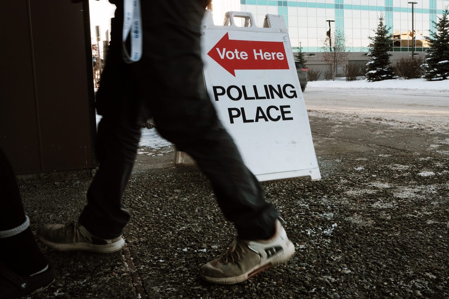 A close-up image shows a "Polling Place" sign with a red arrow and the words "Vote Here" on a sidewalk, directing voters to the entrance of a voting location. Two people are walking by, with only their legs visible. The ground is covered in patches of snow, indicating cold weather, and a modern office building is visible in the background. The image captures the atmosphere of an election day in a wintry setting.