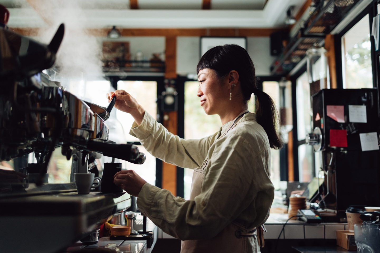 Side view of happy female barista using a coffee maker to heat milk in a jug while standing in a retro cafeteria.