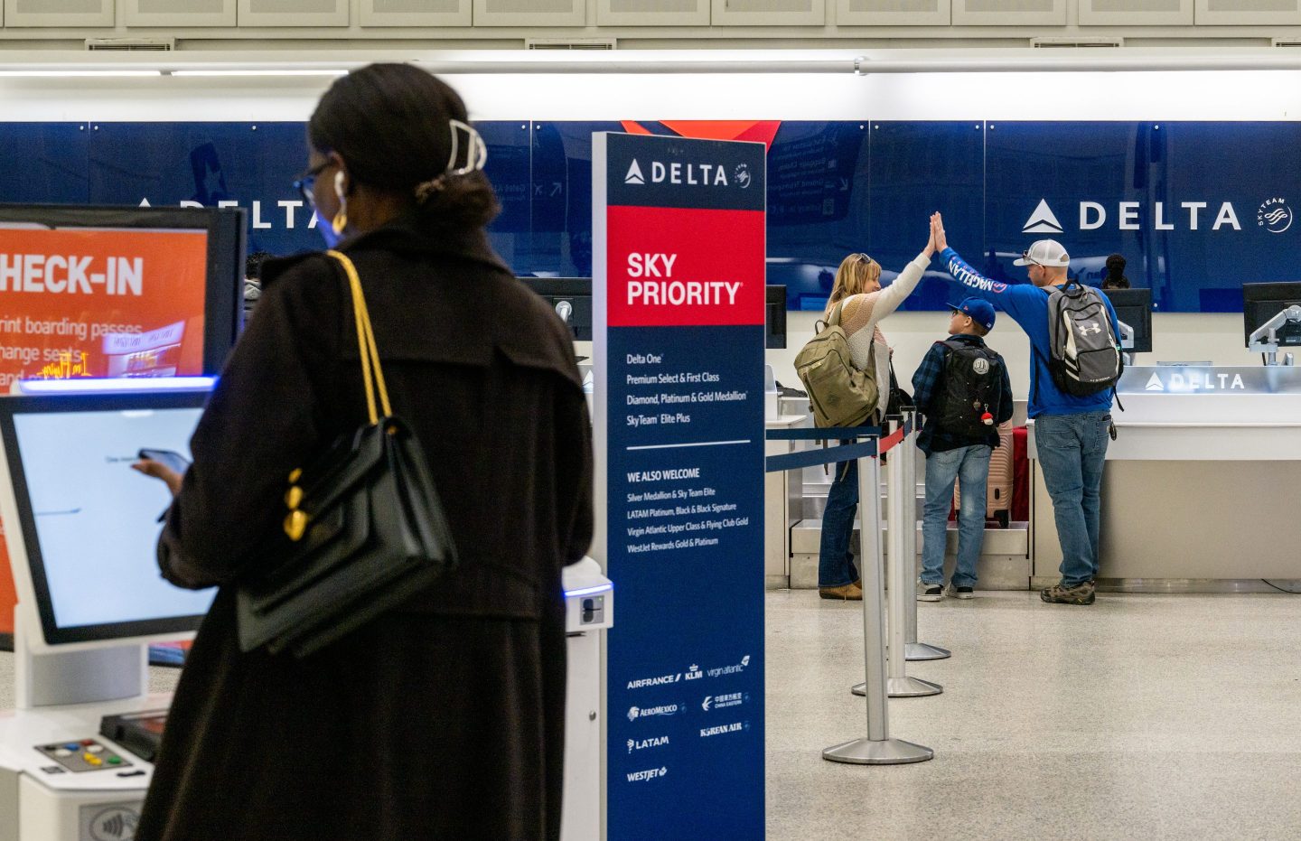 A Delta Airlines check-in area at an airport, featuring a "Sky Priority" sign listing various elite membership tiers for passengers. In the foreground, a woman with a black coat and a large handbag is using a check-in kiosk, while in the background, a family is seen giving high-fives near the Delta counter. The area is organized with stanchions guiding passengers through the check-in process.