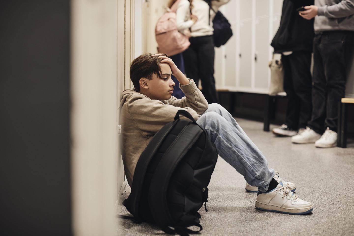 A young teenager sits on the floor near a row of lockers with his backpack next to him