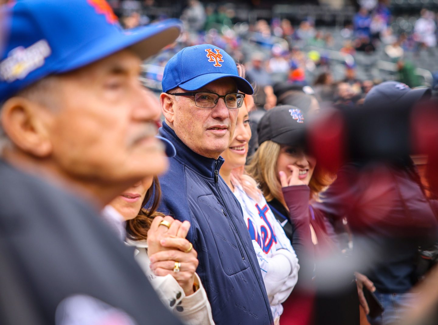 Steve Cohen, who owns the Mets, is seen at an opening game decked out in gear including a baseball cap and vest. He is looking toward the camera wearing glasses.