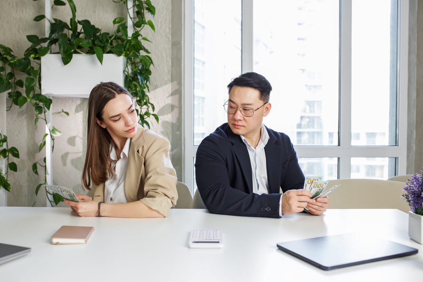 A woman and a man sit at a table in a workplace setting, both holding stacks of money. The woman looks at the man's larger stack of cash with a subtle expression of concern, while the man glances at her, representing the concept of the gender pay gap and unequal earnings between men and women