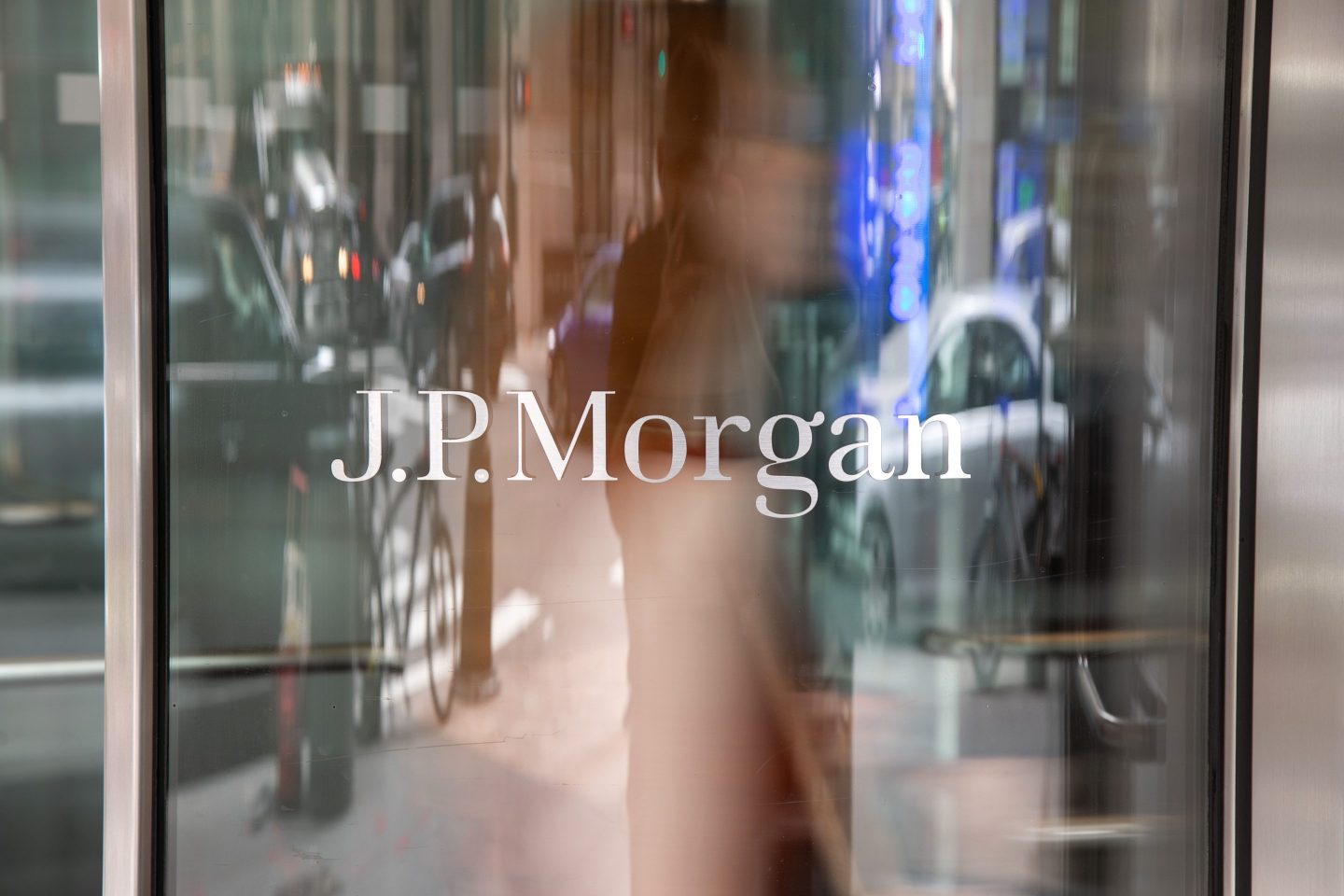 The image shows the entrance of a building with the logo "J.P. Morgan" clearly visible on a glass door. The reflection of street traffic and blurred figures passing by creates a dynamic and bustling urban atmosphere, emphasizing the company's prominent location and activity.