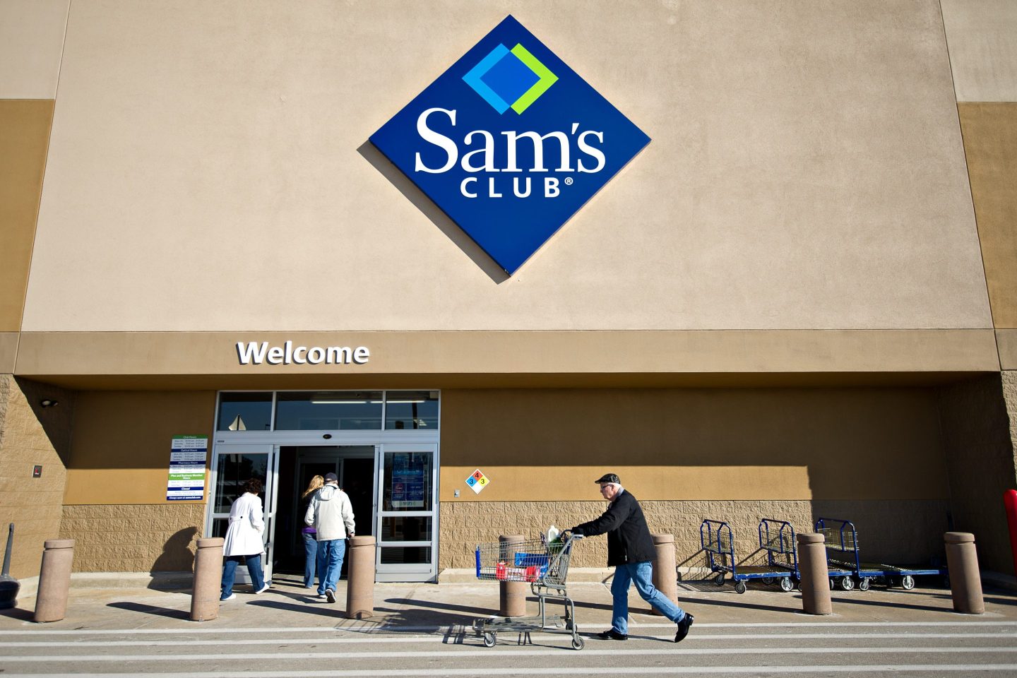 An exterior shot of a Sam's Club store with the logo visibly showing above the sliding door entrance. A shopper is seen pushing their cart beneath the store sign and two other shoppers are near the entrance.