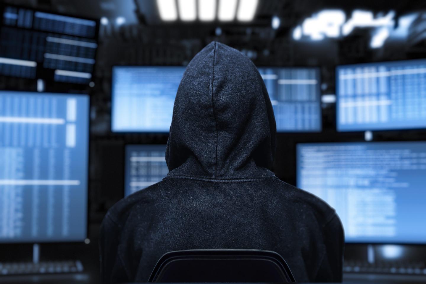 An anonymous hacker in a dark hoodie sits in front of multiple computer screens filled with data. The dimly lit room and the anonymous figure create a sense of secrecy.
