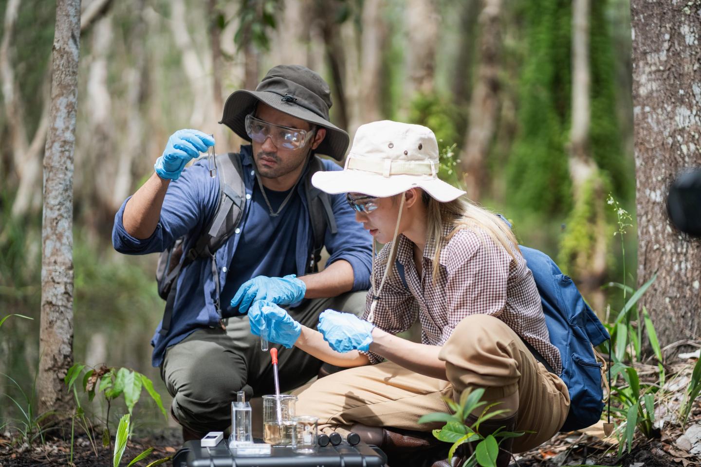 Two researchers observe test tubes while in a wooded area, apparently observing water samples. They are in outdoor safari-like clothing.