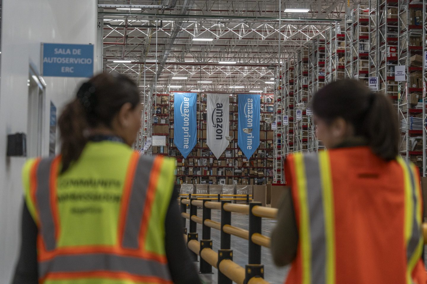 Two blurred Amazon workers stand in the foreground looking away from the camera and toward the open sprawl of an Amazon warehouse. In the background we clearly see Amazon and Amazon Prime flags with logos hanging in the warehouse.