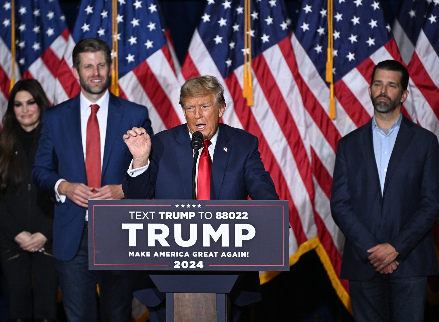 Former President Donald Trump standing onstage behind a lectern with his two adult sons Donald Trump Jr. and Eric Trump by his side.