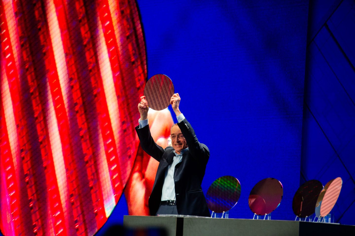 Pat Gelsinger, dressed in a suit on stage, lifts shiny circular computer chips above his head.