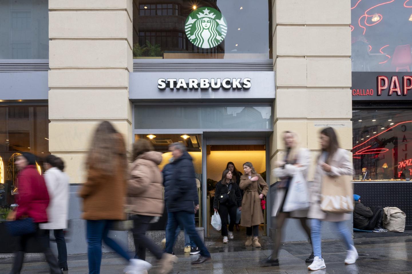 The image shows a busy Starbucks storefront with a crowd of people walking past in both directions. The familiar Starbucks logo with the mermaid is prominently displayed above the entrance. Some people are exiting the store while others are walking by on the street, with a mix of blurred motion capturing the bustling urban environment. The setting appears to be in a city, with a nearby brightly lit shop visible in the background.