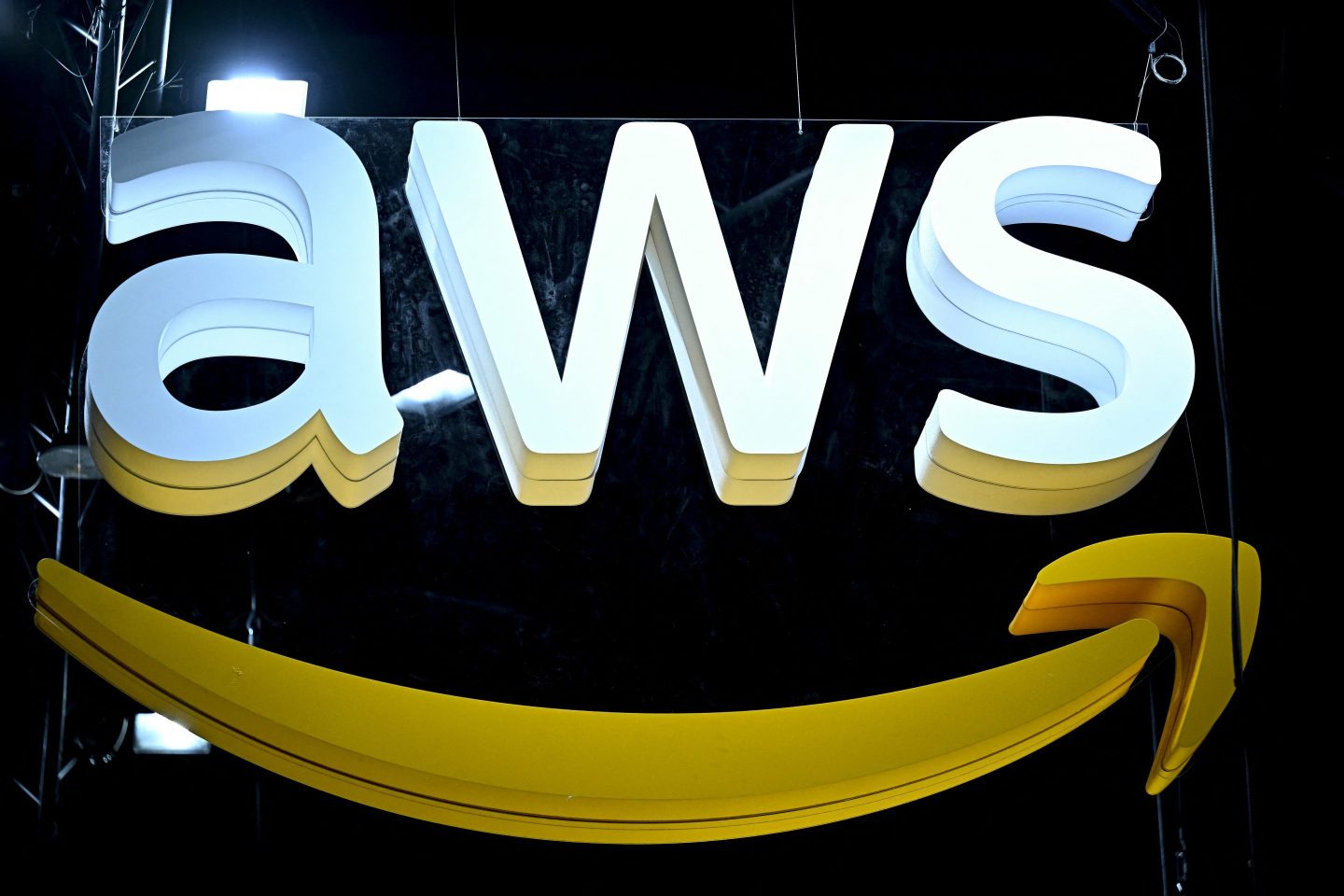 The image shows a large 3D "AWS" logo, which stands for Amazon Web Services. The logo is prominently displayed with bold white letters and a swooping yellow arrow underneath, resembling the Amazon logo. The sign is set against a dark background, and the lighting above the logo adds a dramatic effect to its appearance.